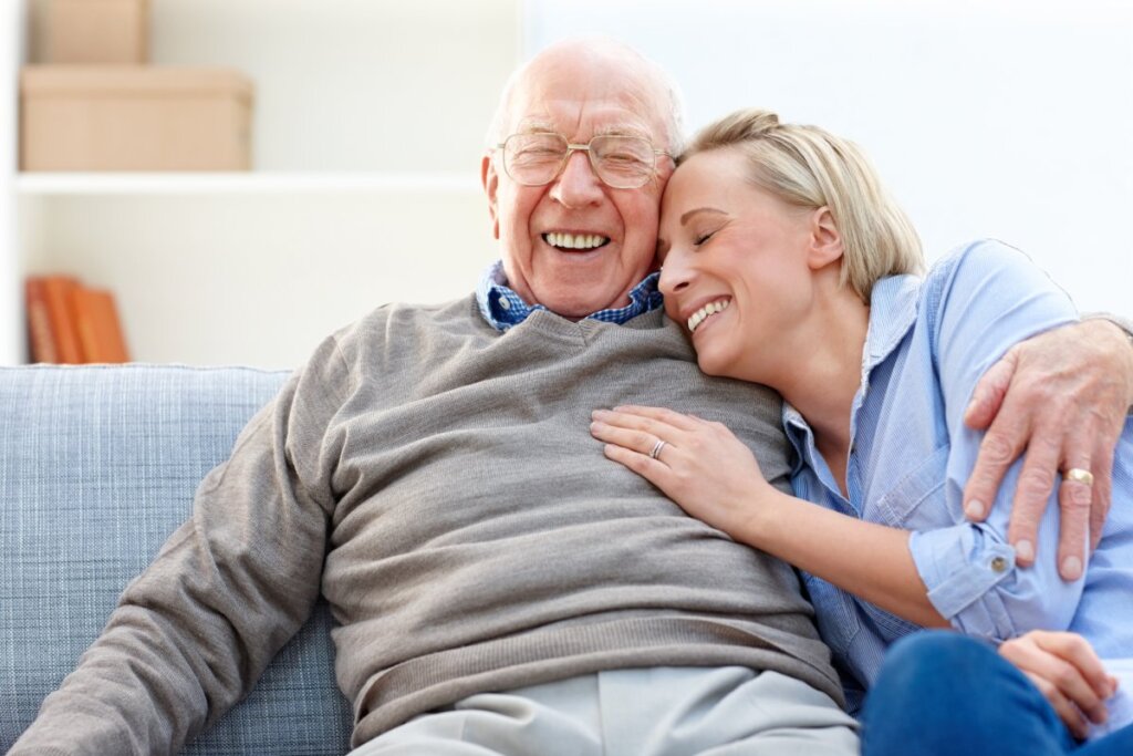 A close-up of an elderly man and woman sitting on the couch hugging, the woman has her hand on the mans chest.