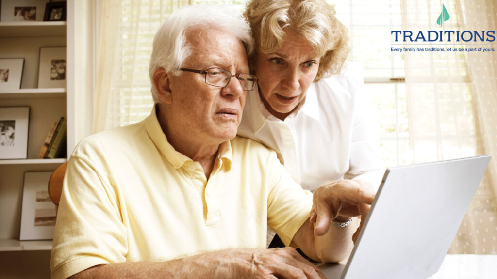 a confused elderly man with grey hair with a concerned-looking elderly woman with blonde curly hair looking at a laptop screen at their home office with Traditions logo on the top right corner