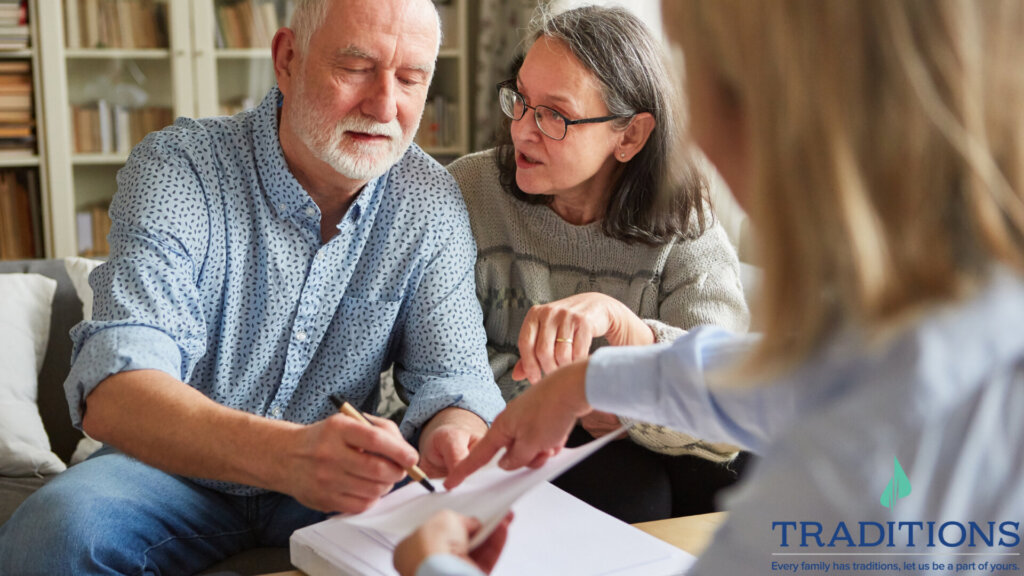 A close up of an elderly couple sitting on a couch with a woman with blonde hair sitting across from them holding up a piece of paperwork over a pile of paper. The woman with blonde hair is pointing down at the power and the elderly man has a pen up to the power with the elderly woman looking at him. Traditions logo at the bottom right corner.