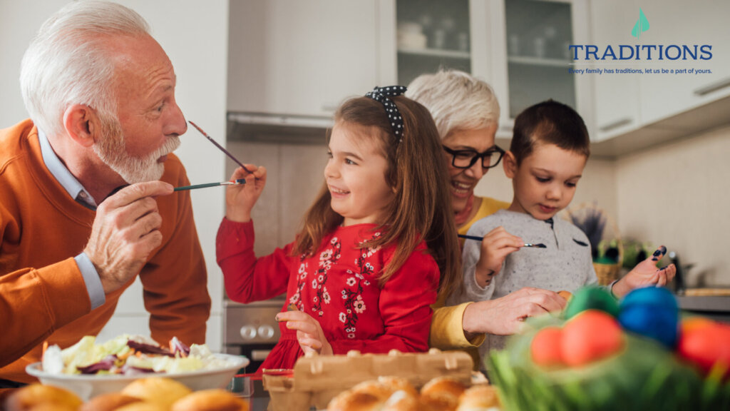 An elderly couple is painting with kids. The young girl is painting the old man's nose. Traditions logo at the top right corner