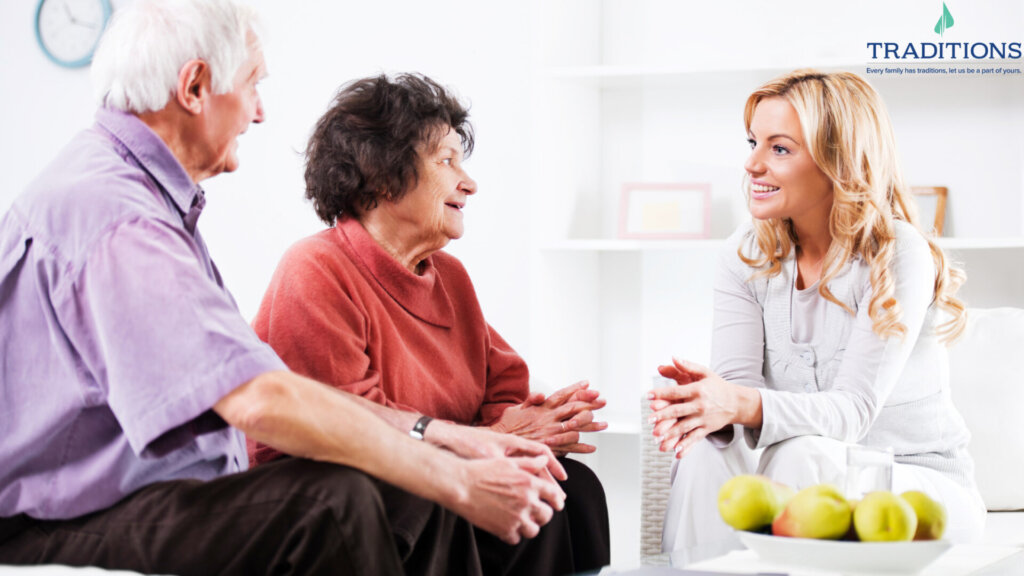 a young blonde woman talking to an elderly couple in a living room with Traditions logo on the top right corner