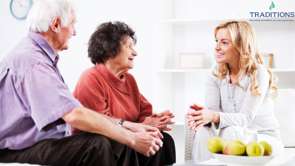 An elderly couple sitting on a couch speaking to a young woman with blonde hair sitting in a car. Traditions Logo in the top right corner.