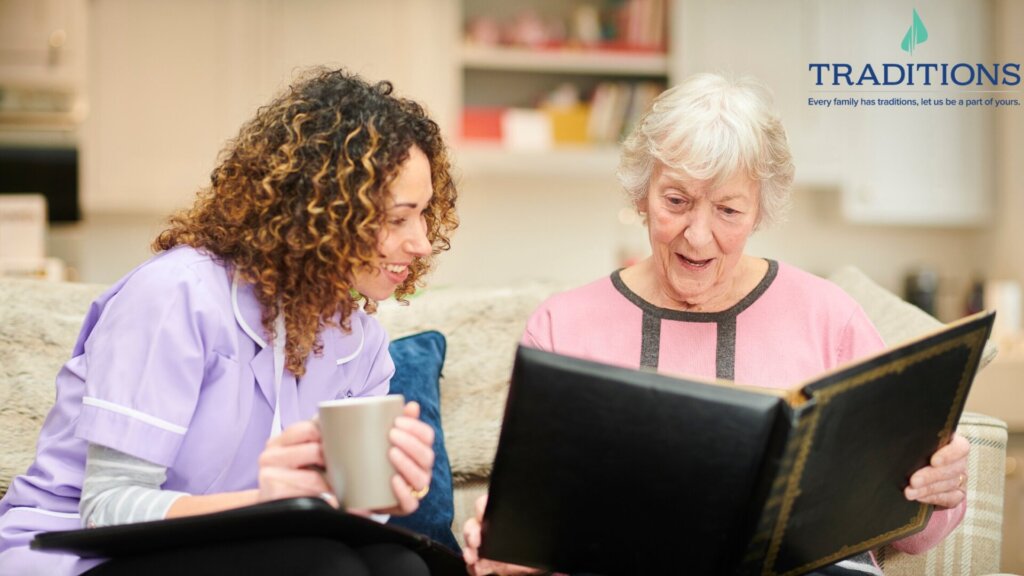 Photo of an elderly woman with white hair looking at an old photo album and showing it to a younger woman with curly brown hair who is holding a coffee mug