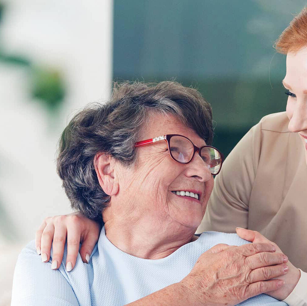 A close-up of an older woman wearing red glasses is holding hands with a caregiver wearing tan scrubs behind her.