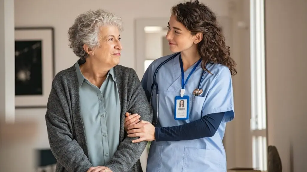 A female caregiver wearing blue scrubs walks down the hall with an older woman wearing a blouse and a cardigan. The caregiver has her hands around the woman's arm.