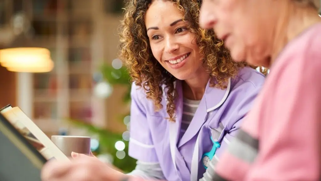 A female caregiver wearing purple scrubs and an older woman wearing a pink shirt sit together on a couch, looking at a photo album.