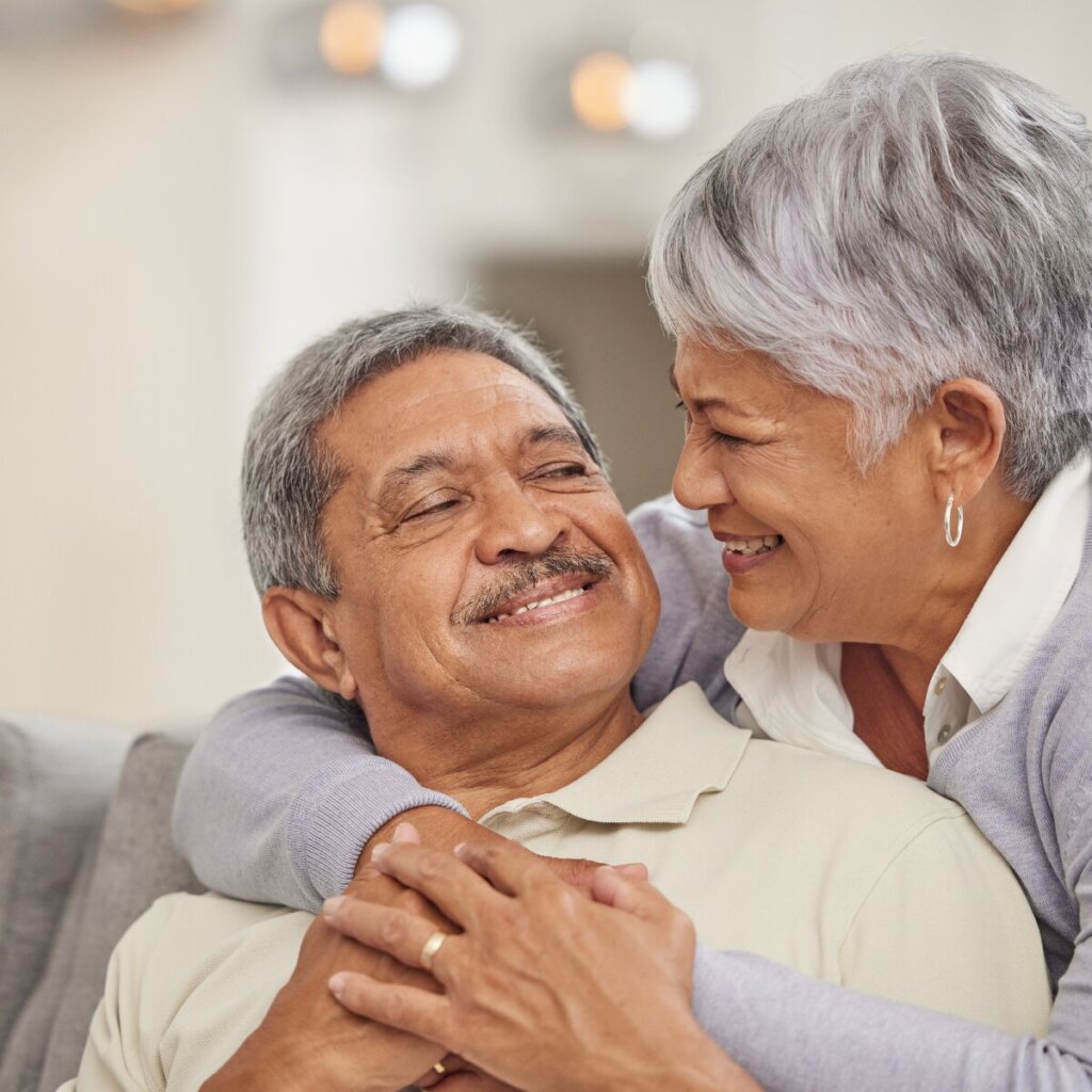 An older couple posing together on a gray couch; the woman has her hands around the man’s shoulder, both smiling.