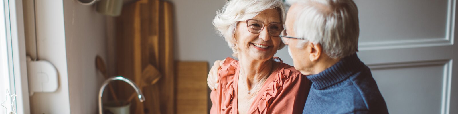 An older couple posing together in a kitchen; the man has his hand on the woman’s shoulders, and they are both smiling.