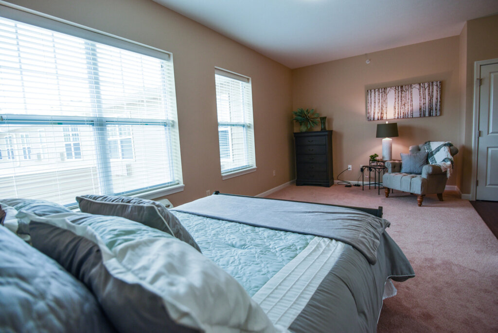 bedroom at Traditions of Beavercreek featuring a bed near large windows, with a dresser and cushioned chair