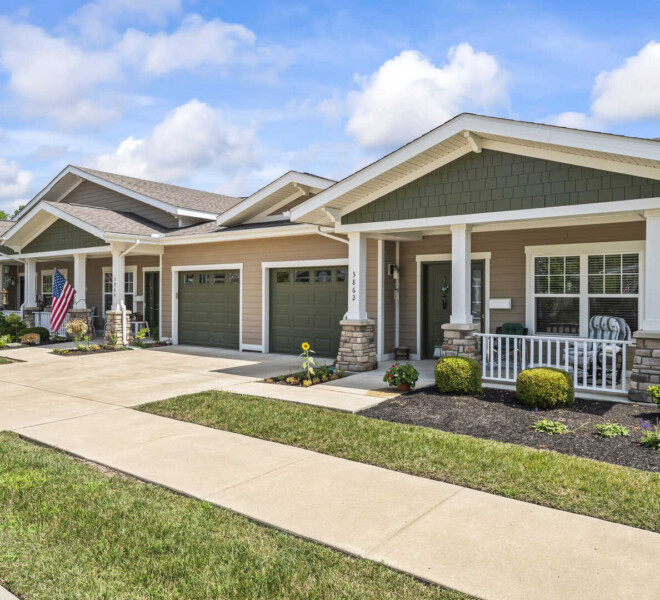 Front view of a row of garden homes at The Traditions of Beavercreek retirement community in Ohio, featuring front porches, attached garages, American flags, manicured landscaping, and a concrete sidewalk