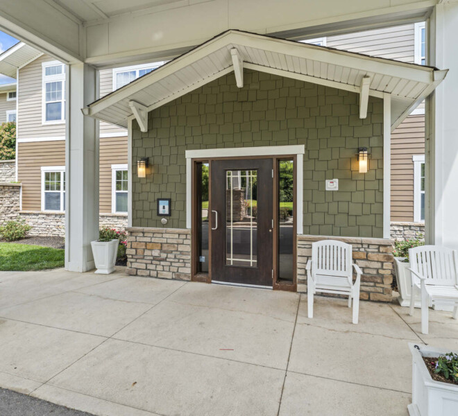 Close-up of the main entrance doors at The Traditions of Beavercreek retirement community in Ohio, with stone accents, white chairs, and potted plants.