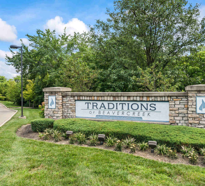 Entrance sign for The Traditions of Beavercreek retirement community in Ohio, surrounded by neatly trimmed bushes, flowering plants, and a backdrop of trees