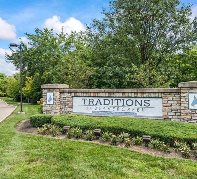 Entrance sign for The Traditions of Beavercreek retirement community in Ohio, surrounded by neatly trimmed bushes, flowering plants, and a backdrop of trees